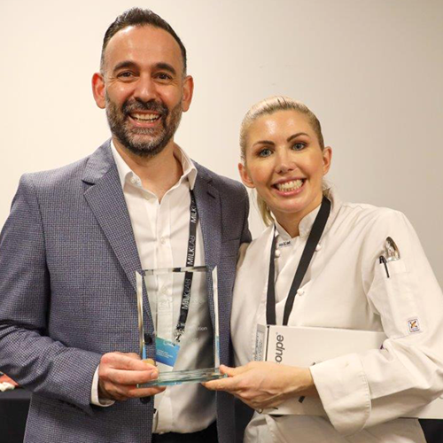 A woman in chef's jacket smiling, receiving a rectangular glass trophy from a smiling man. 