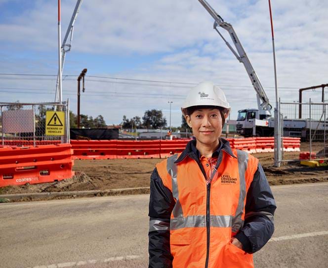 Woman wearing level high visibility jacket - that has "level crossing removal" printed on the front - standing in front of the worksite with heavy machinery in background.