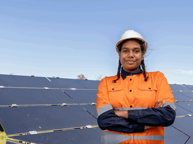 Smiling woman standing outside in safety work clothing and helmet with her arms crossed in front of solar panels.